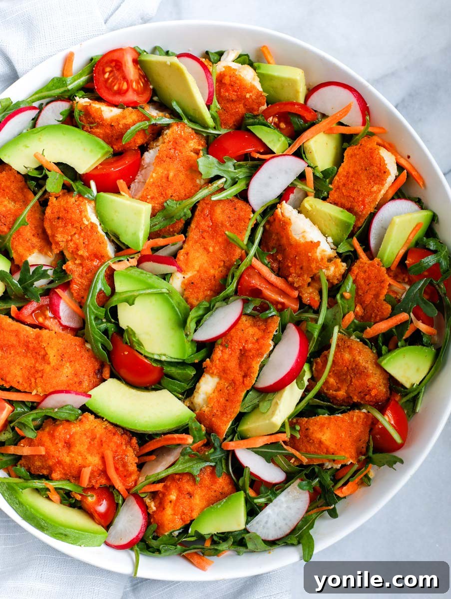 Arugula and other fresh vegetables topped with spicy buffalo chicken pieces in a white bowl, ready for dressing.