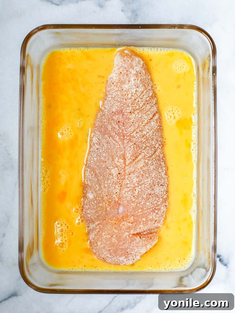 Raw chicken breast being dipped into an egg wash mixture.