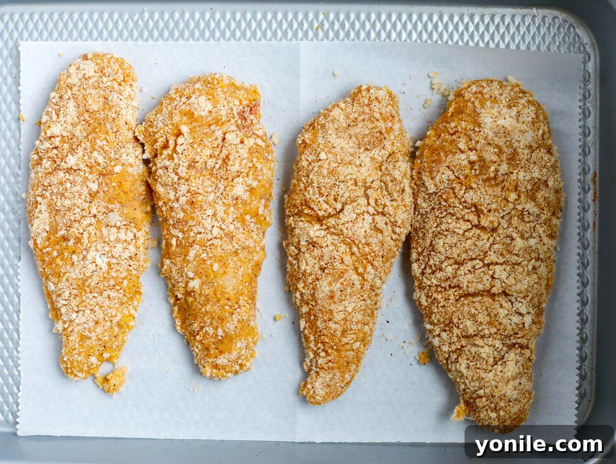 Four breaded chicken breasts neatly arranged on a parchment-lined baking tray, ready for the oven.