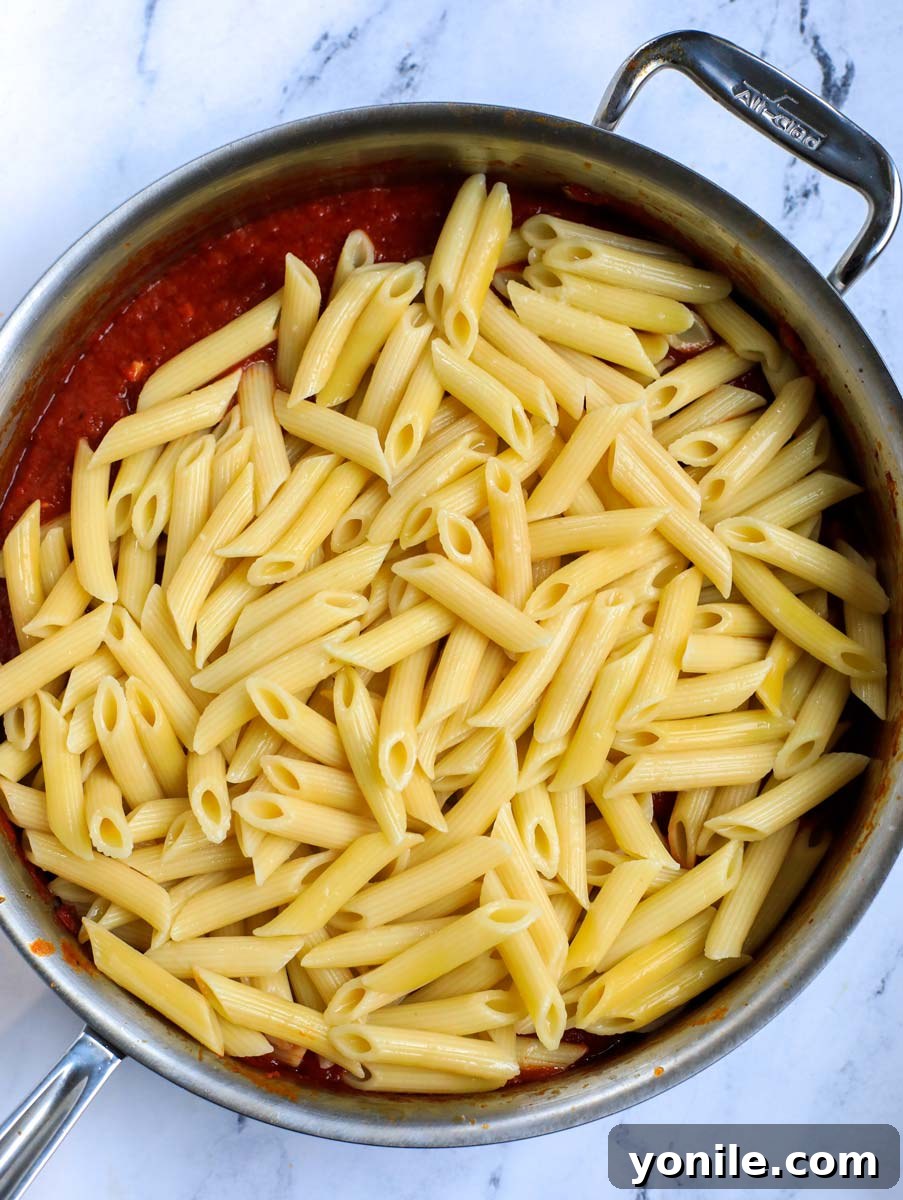 Penne pasta being added to the rich tomato sauce in a pot.