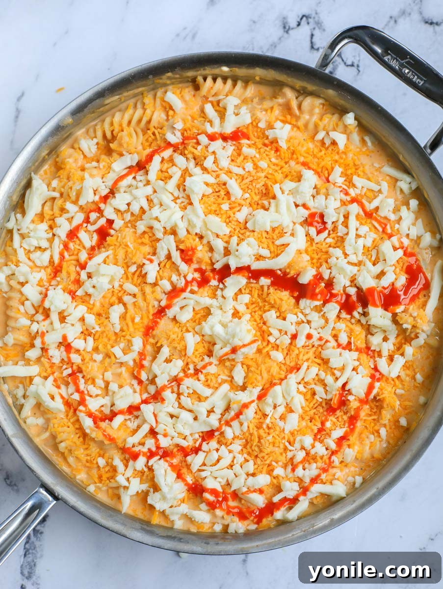 Additional shredded cheese and a drizzle of buffalo sauce layered on top of the pasta mixture in a pot, ready for baking.