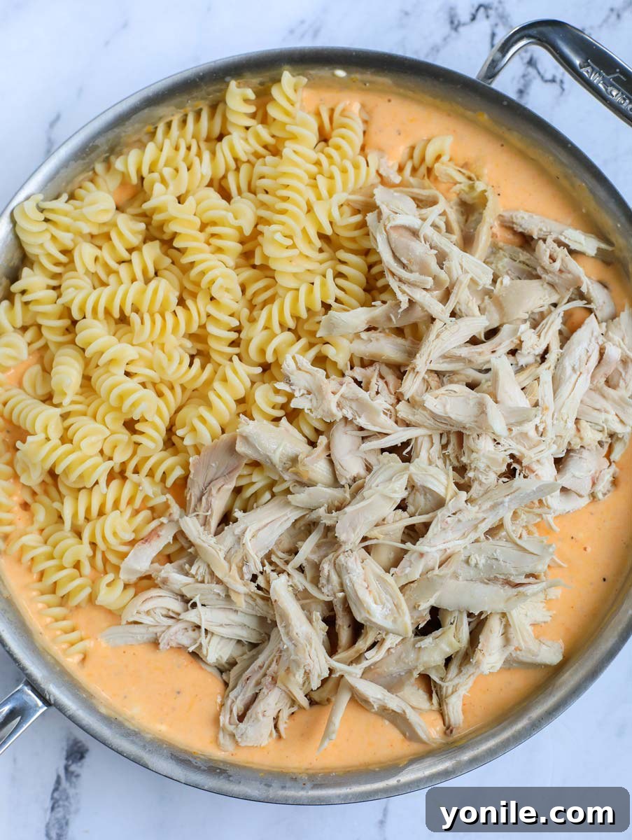 Cooked pasta and shredded chicken being added to the creamy cheese sauce in a large pot.