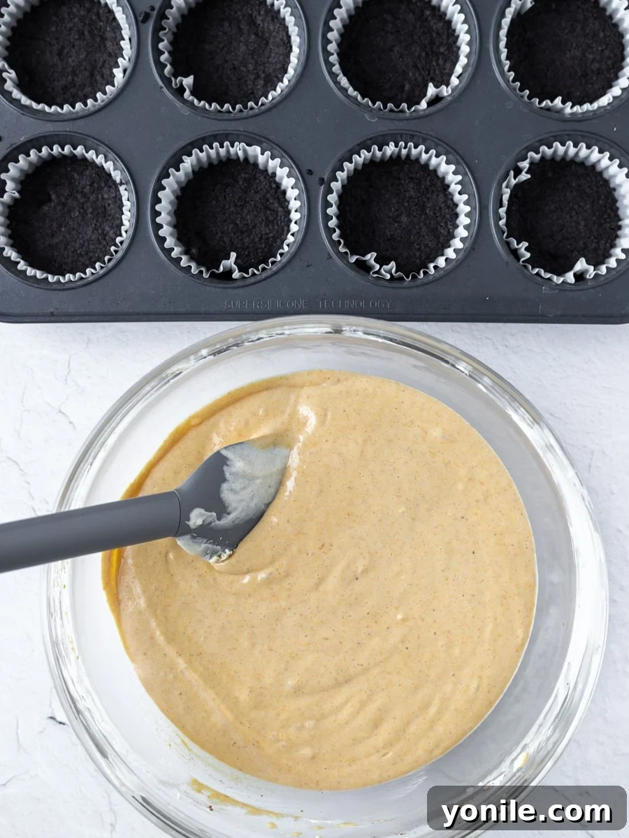 Overhead shot of creamy pumpkin cheesecake filling mixture in a mixing bowl next to a muffin tin with pre-baked Oreo cookie bases in molds.