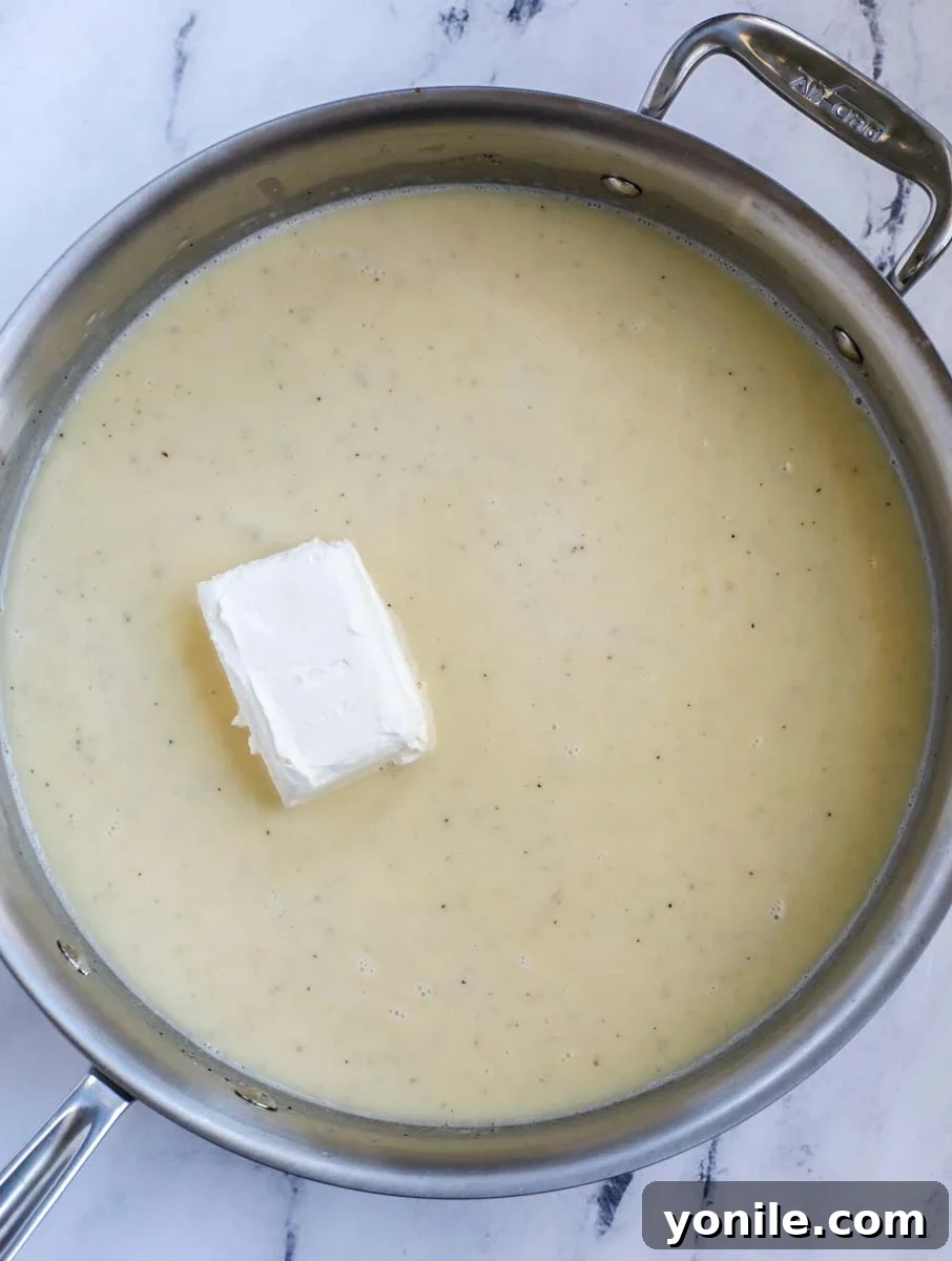 A stainless steel skillet containing a creamy, light-colored casserole sauce with a block of softened cream cheese being stirred in.
