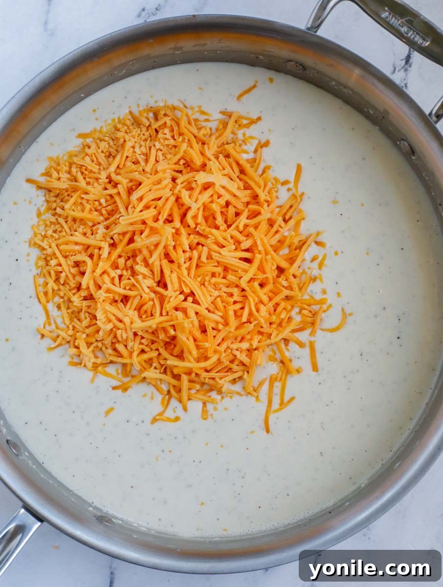 A silver skillet with a creamy sauce and a generous pile of shredded sharp cheddar cheese, about to be mixed in.