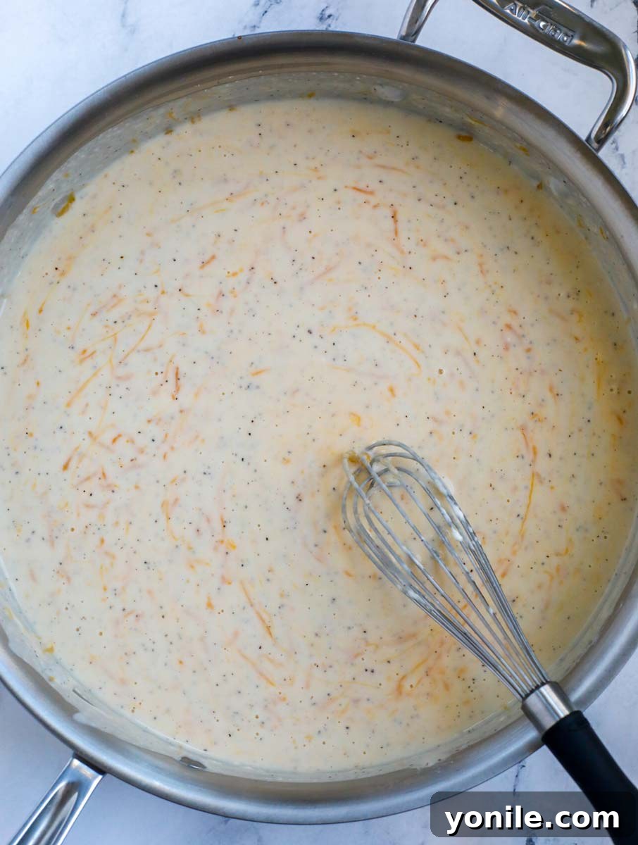 A silver skillet filled with a creamy, cheesy casserole sauce, with a wire whisk resting on the right side, showing the smooth texture.