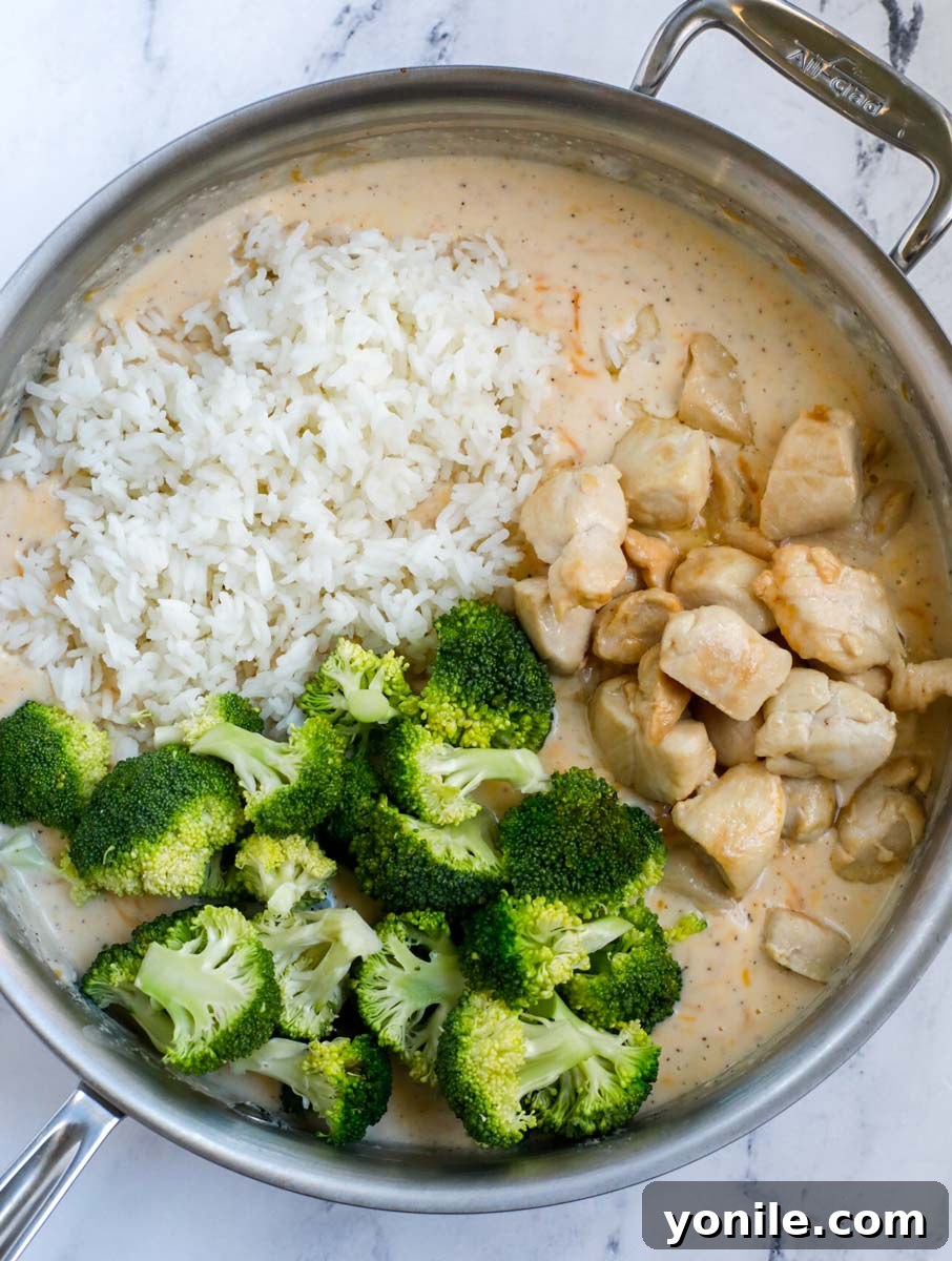 A silver skillet with a smooth, creamy casserole sauce at the bottom, and layers of cooked rice, chicken, and broccoli placed on top, ready to be combined.