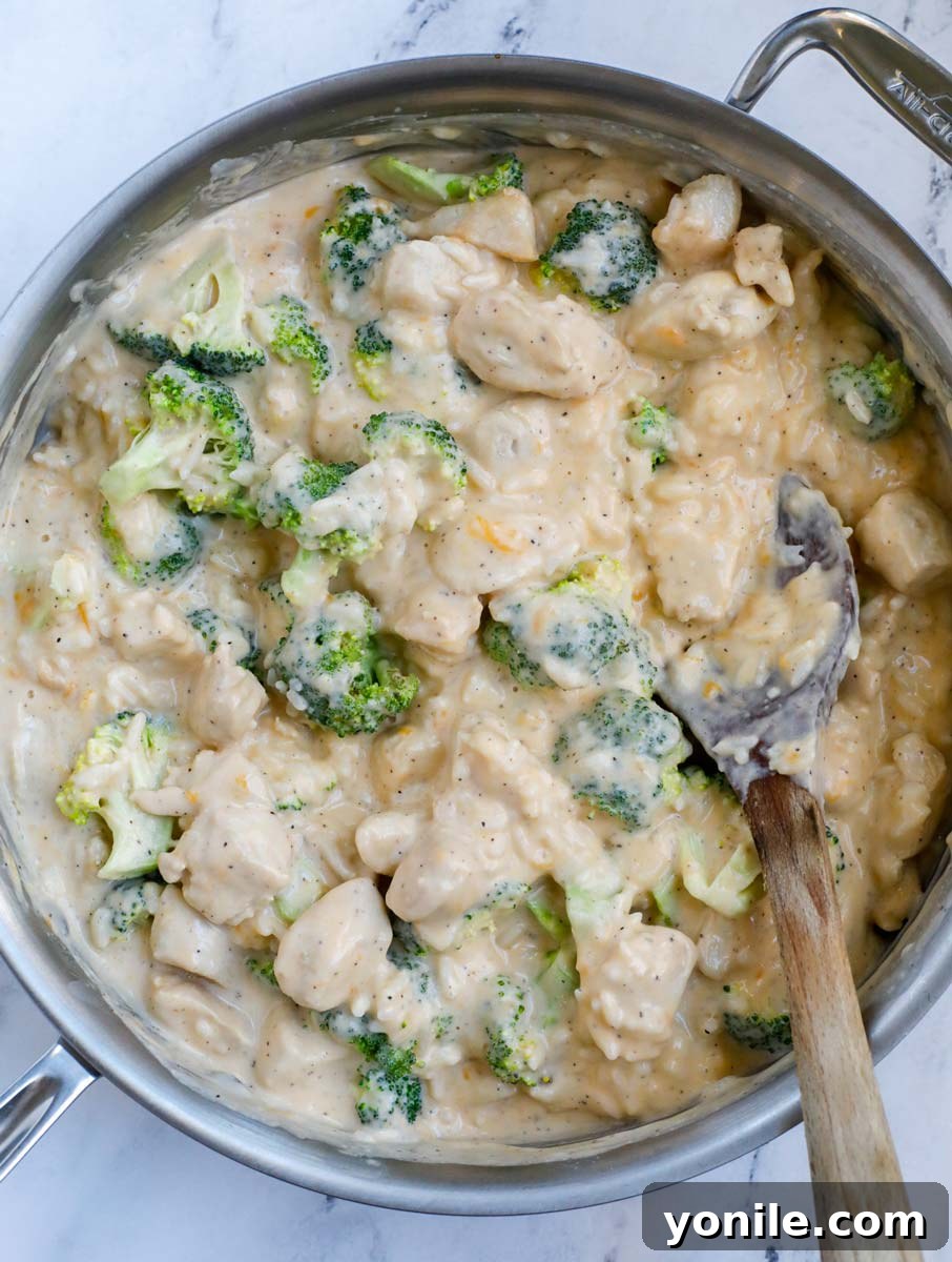 A silver skillet filled with a perfectly mixed blend of chicken, broccoli, and rice, all coated in a creamy sauce, with a wooden spoon resting on a white counter.