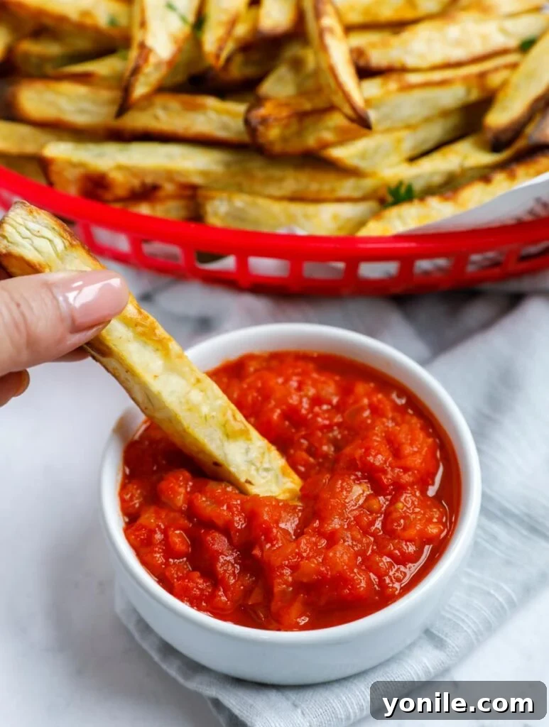 A hand dipping a crispy sweet potato fry into a bowl of homemade tomato sauce.
