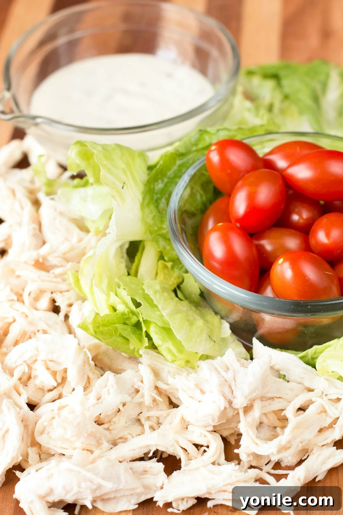 Effortless Chicken Caesar Wraps 4 Chicken Caesar Wrap Ingredients laid out on a cutting board, including chicken, lettuce, tomatoes, tortillas, and dressing components.