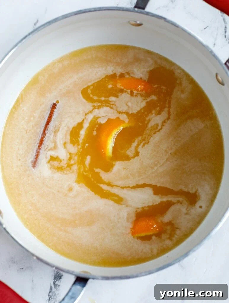 Orange juice, orange peel, sugar, and a cinnamon stick simmering gently in a small saucepan on a stovetop.