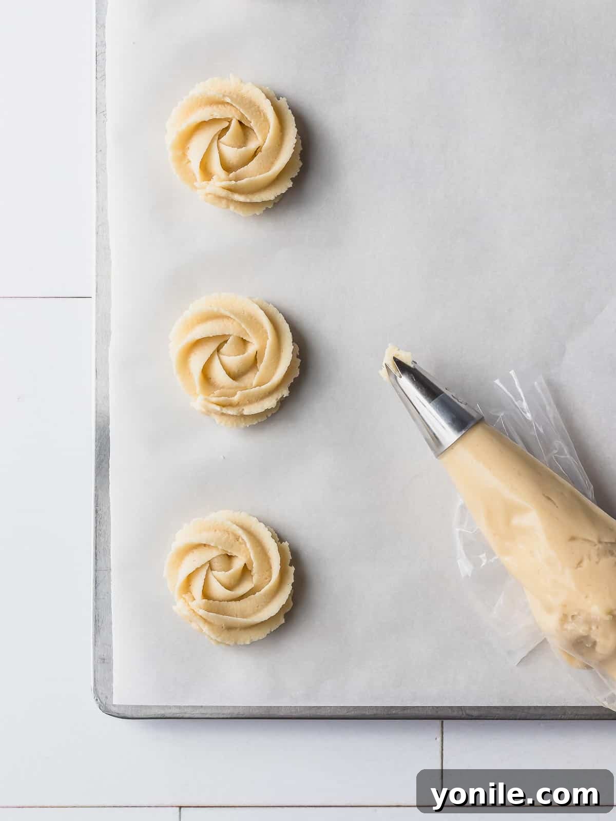 A pastry bag with a star tip actively piping swirls of Danish butter cookie dough onto a baking sheet lined with parchment paper, creating uniform cookie shapes.