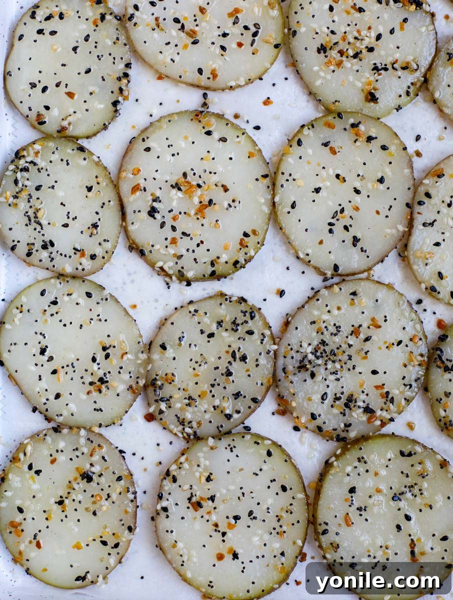 Everything Bagel Chips 5 An overhead shot of numerous thinly sliced potato rounds neatly laid out in a single layer on a parchment-lined baking sheet, glistening with oil and ready for seasoning before baking.