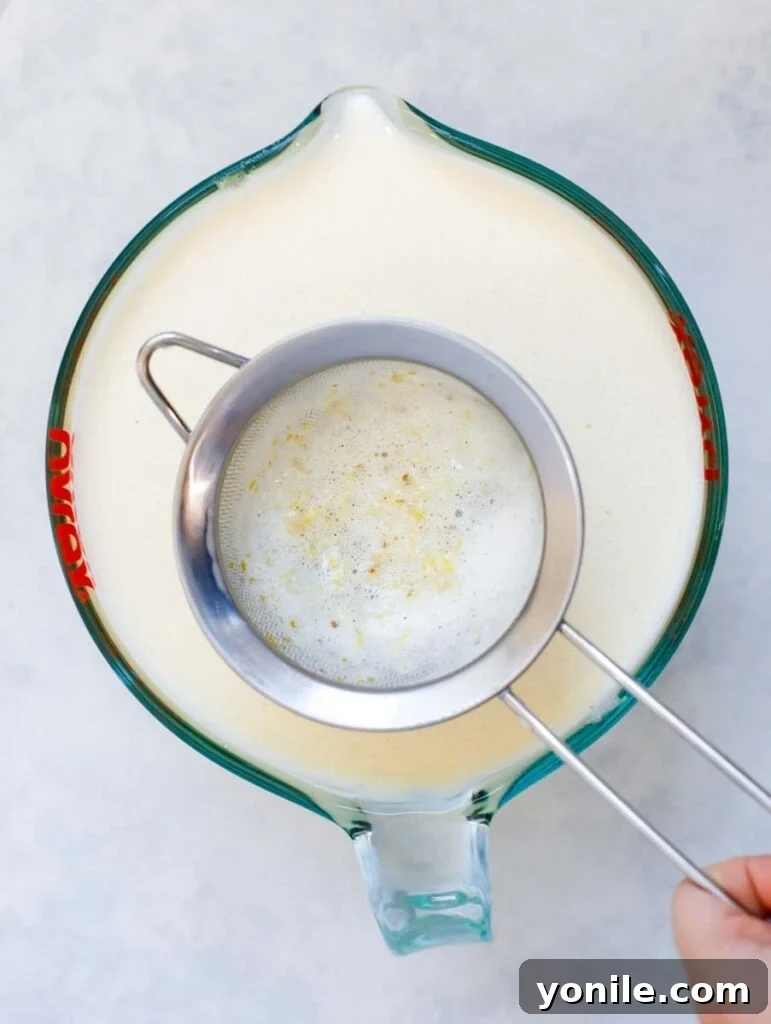 Straining the finished custard mixture through a fine-mesh sieve into a pitcher to remove impurities.