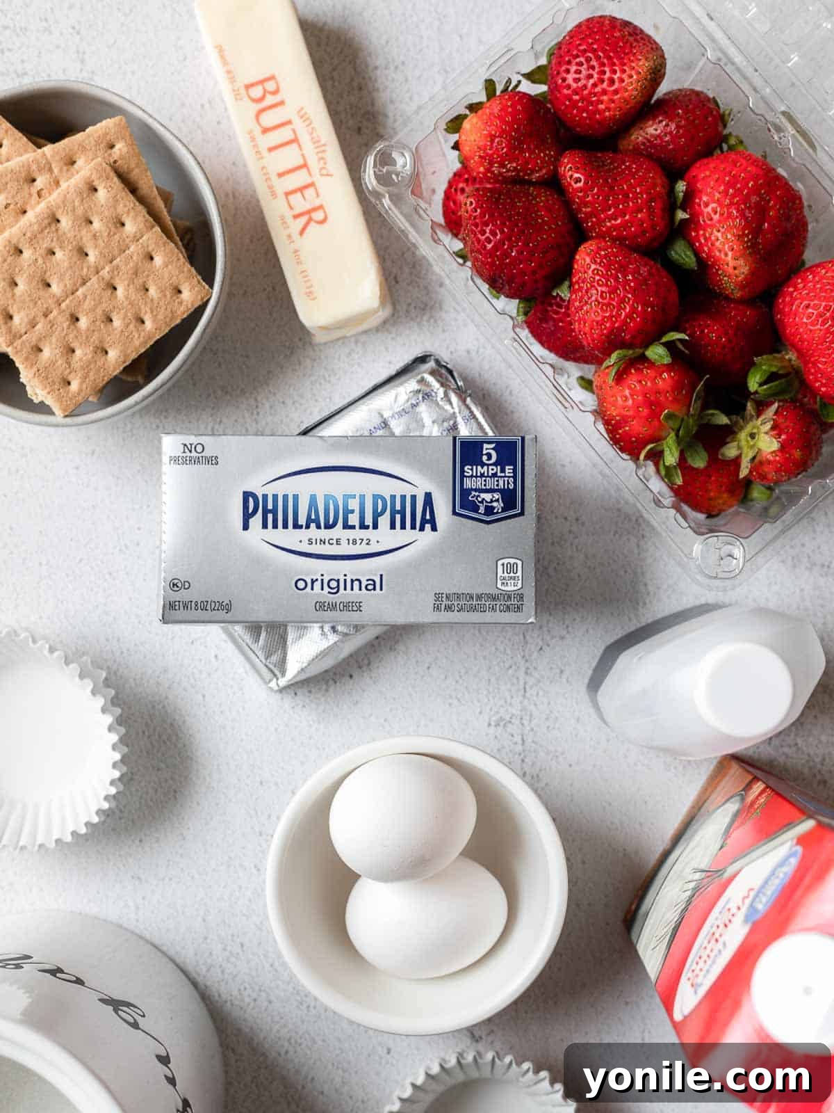 A flat lay of fresh ingredients for mini strawberry cheesecakes, including graham crackers, cream cheese, eggs, strawberries, sugar, and butter.