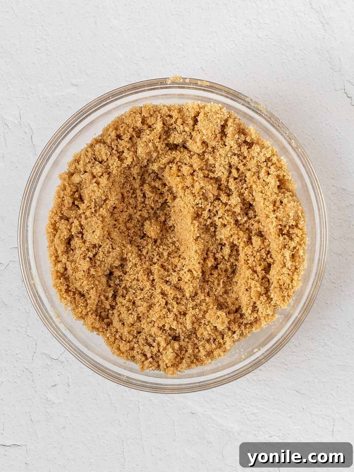 Close-up of graham cracker crumbs being mixed with melted butter and sugar in a glass bowl to form a cheesecake crust.