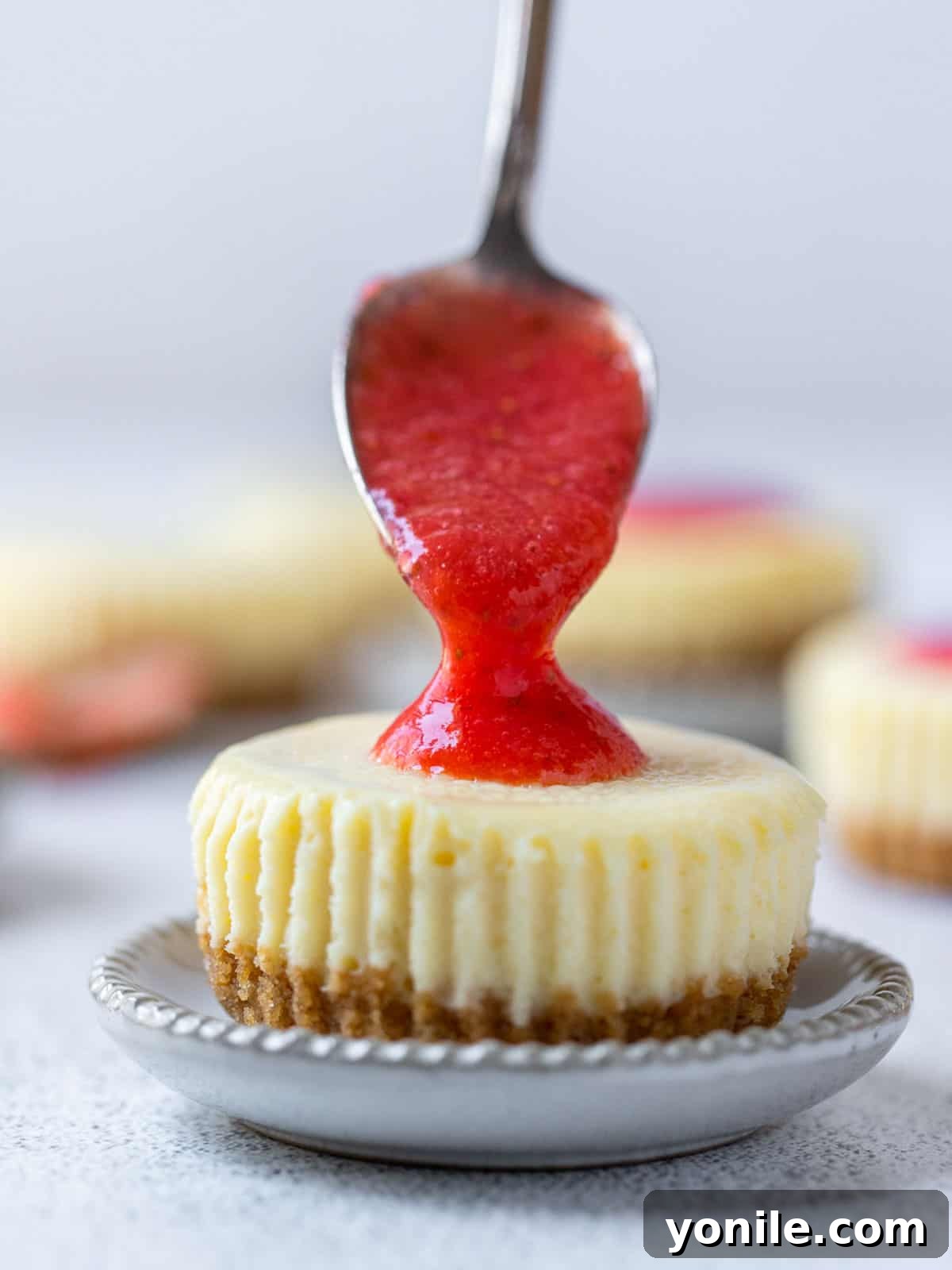 A spoon gently adding fresh strawberry sauce as a topping onto a mini strawberry cheesecake.