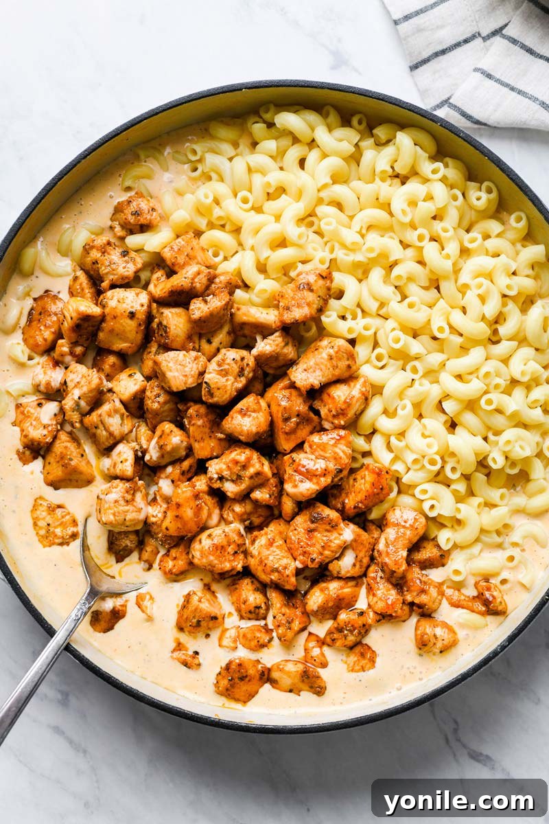 Cooked macaroni and seasoned chicken pieces being stirred into the creamy Cajun cheese sauce in a skillet.