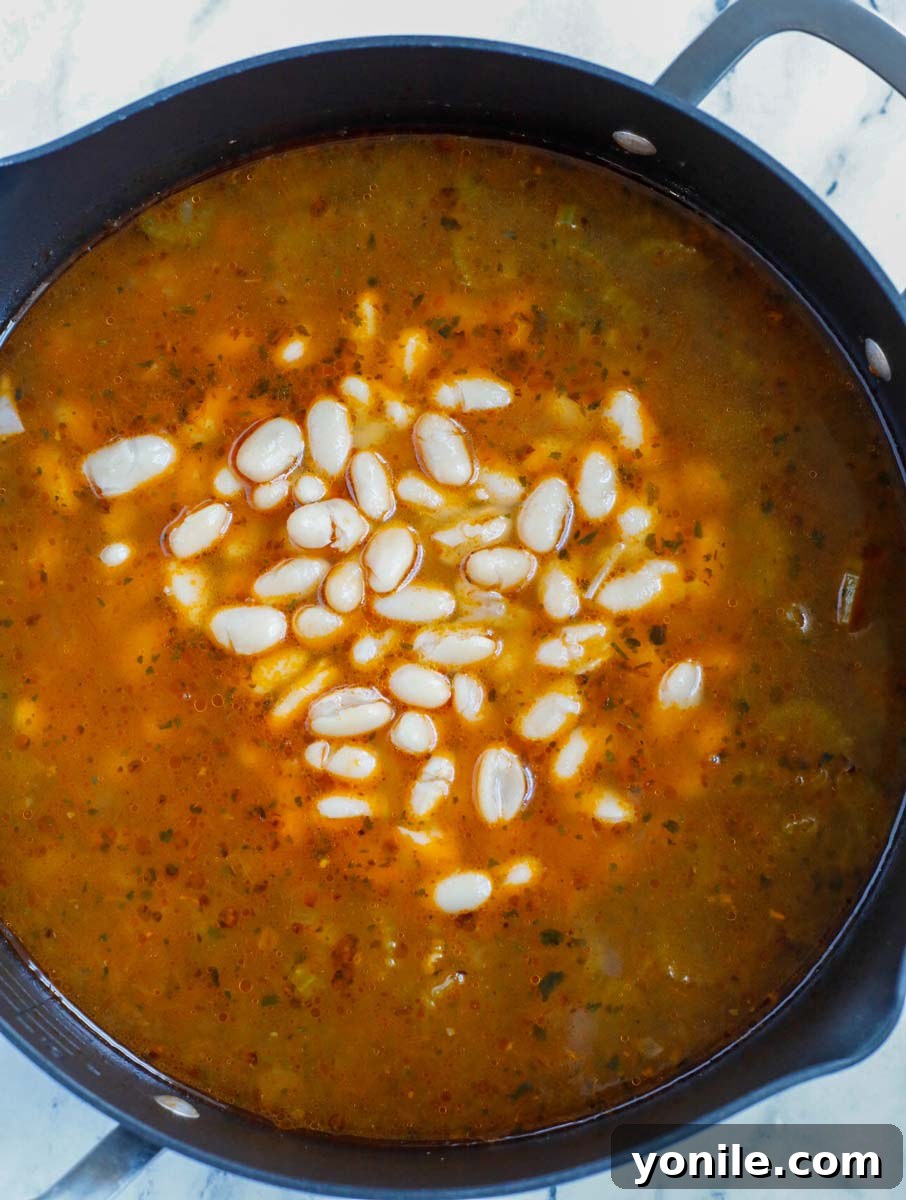 Creamy white cannellini beans being gently poured from a can into a simmering pot of rich Tuscan White Bean Soup, ready to absorb its delicious flavors.