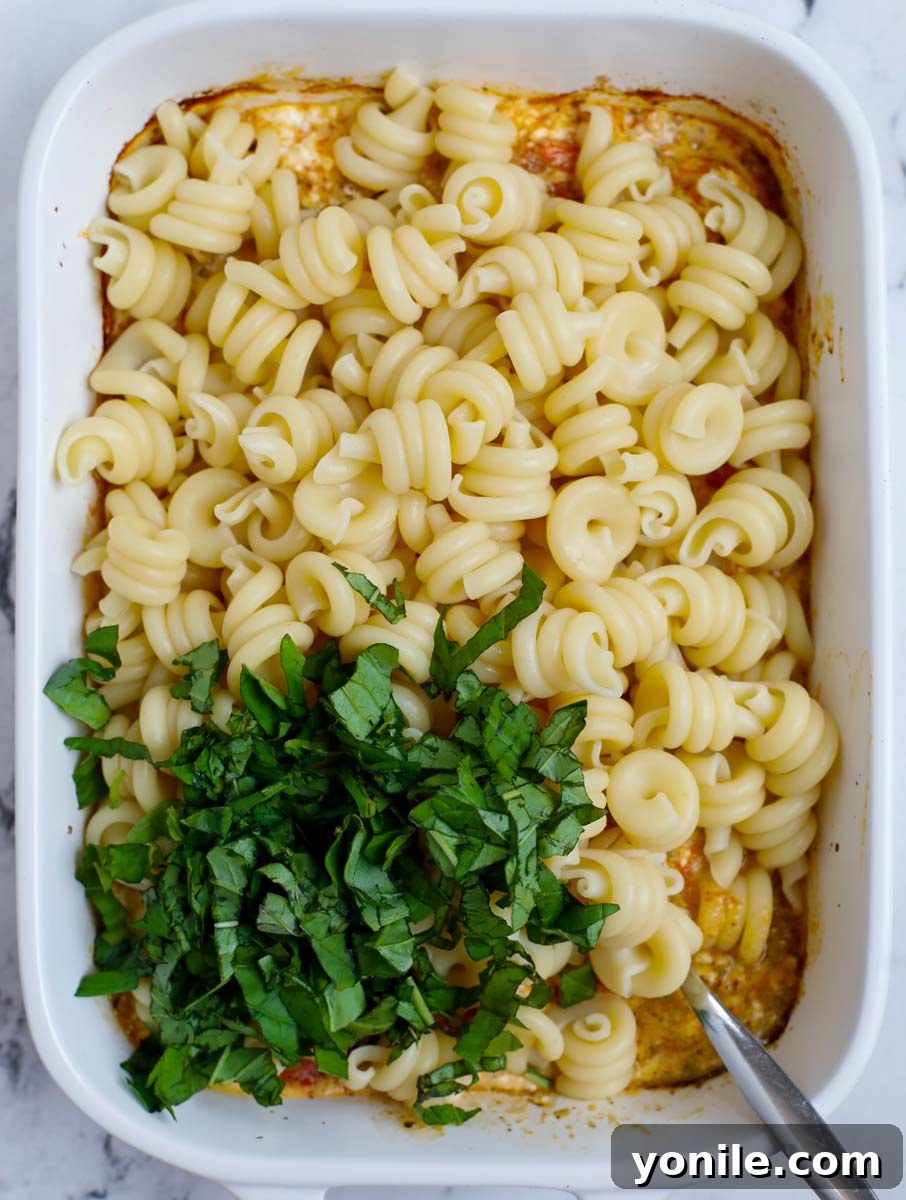 Cooked pasta and fresh basil being added to the creamy feta and tomato sauce in the baking dish, ready to be tossed.
