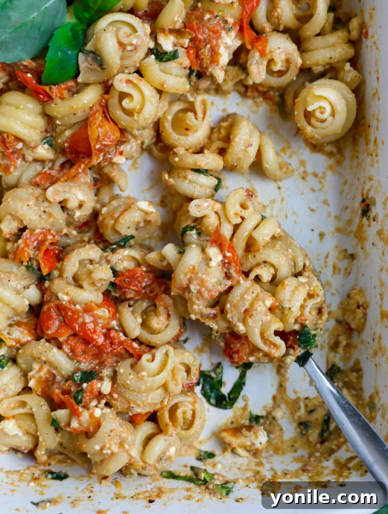 A serving spoon scooping out a generous portion of creamy feta pasta from a white casserole dish, highlighting its rich texture and ease of serving.