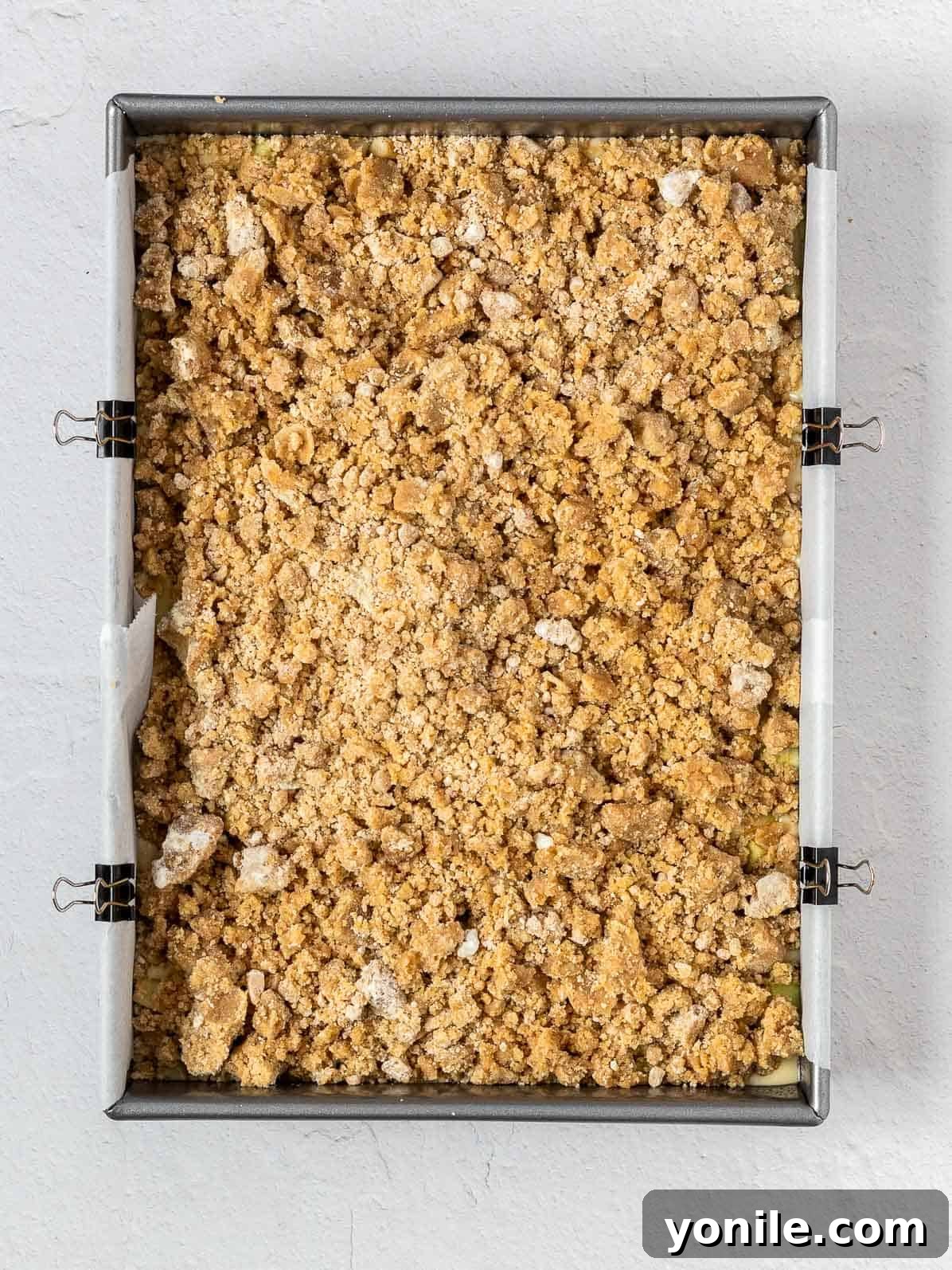 An unbaked apple crumb cake, generously topped with crumble, resting in a rectangular pan before entering the oven.