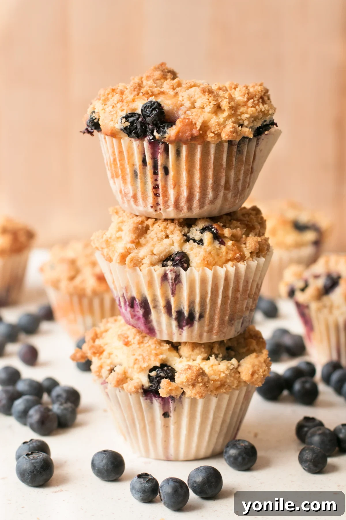 A stack of golden-brown blueberry crumb muffins, glistening with streusel topping, sitting on a white surface and surrounded by loose, vibrant blueberries.