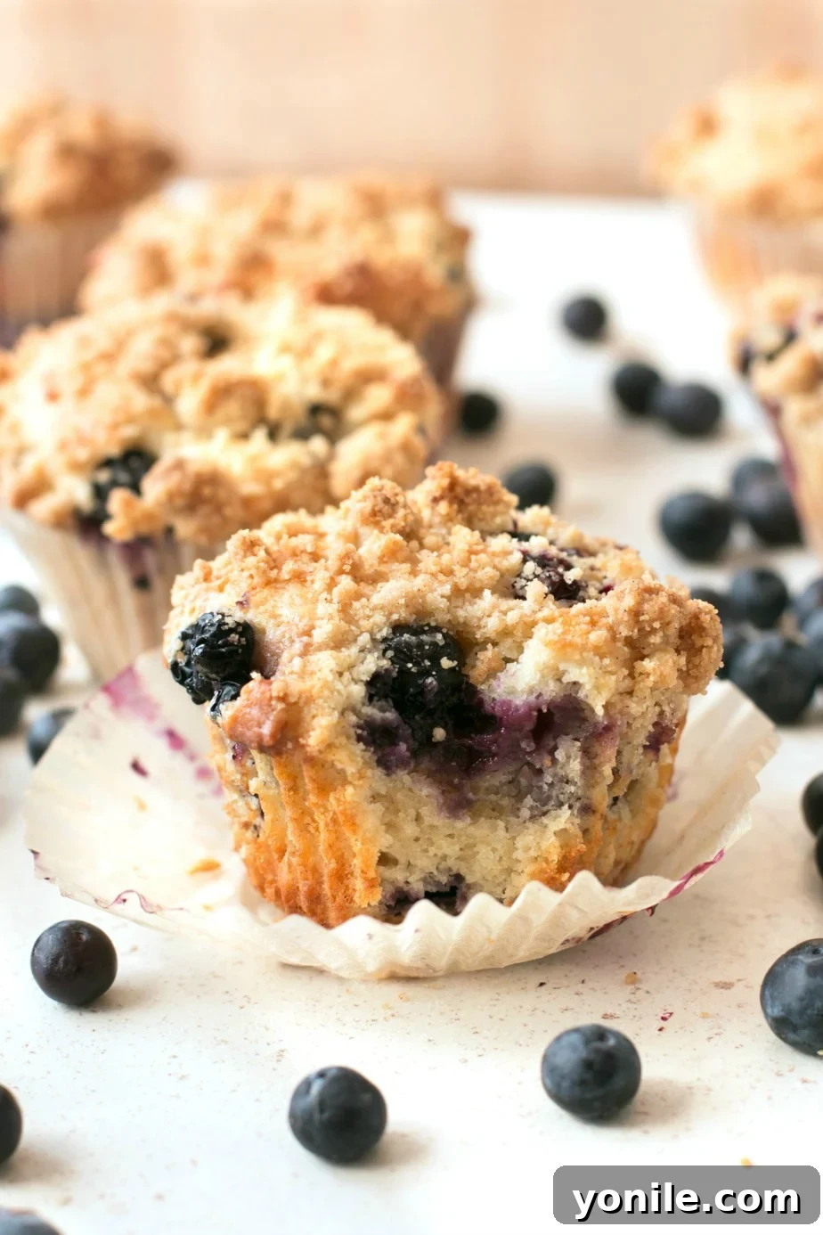 A close-up shot of a plate of blueberry crumb muffins, one with a bite taken, revealing its moist interior and plump blueberries, all on a clean white background.