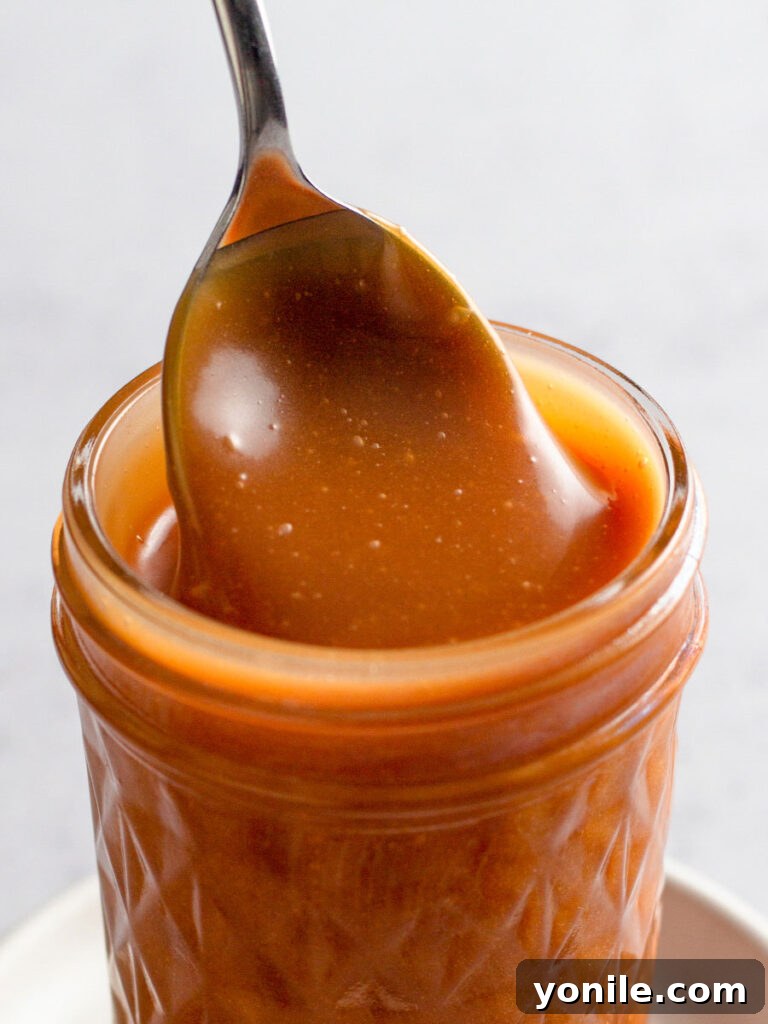 Close up of a glass jar filled with golden homemade caramel sauce, with a spoon resting inside, ready to be drizzled.