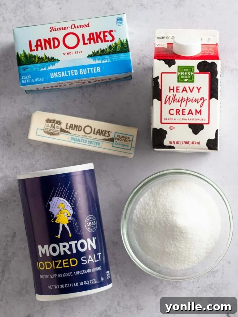 Ingredients for making homemade caramel sauce laid out on a white surface: granulated sugar, cubes of unsalted butter, heavy cream in a measuring cup, and a small pinch of salt.