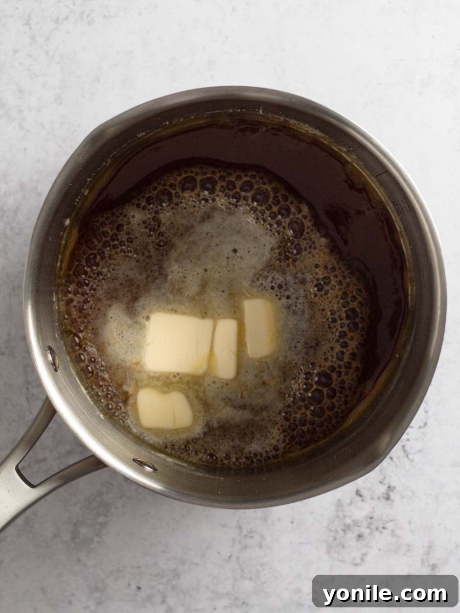 Small cubes of unsalted butter being added to the hot, caramelized sugar in a saucepan, causing a slight bubbling reaction.