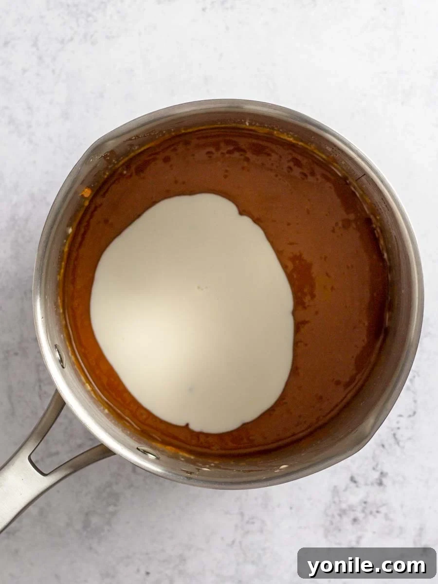 Heavy cream being slowly poured into the butter-caramel mixture in a saucepan, creating a bubbling and frothing effect.