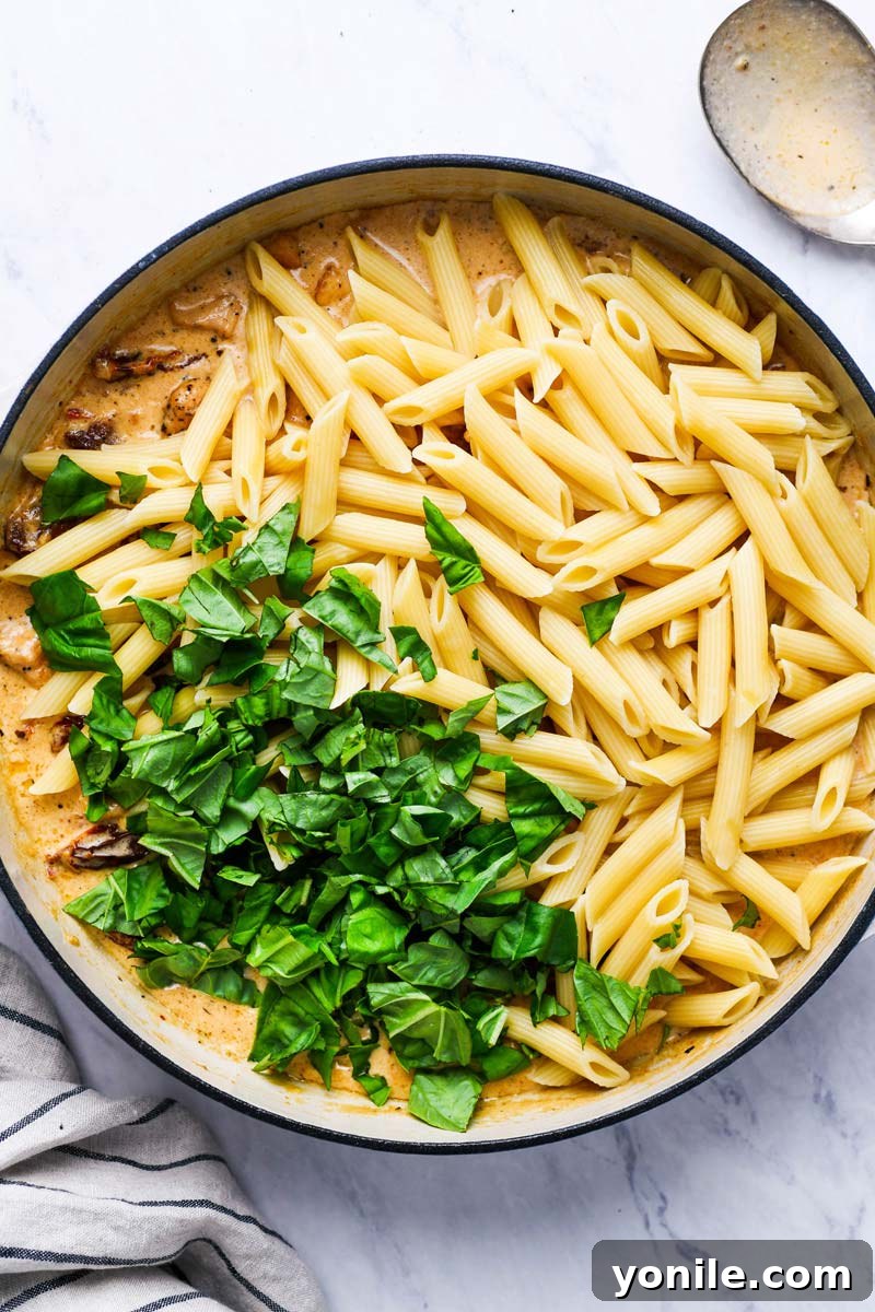 Adding cooked pasta and fresh basil to the cream sauce in a pot to be coated.