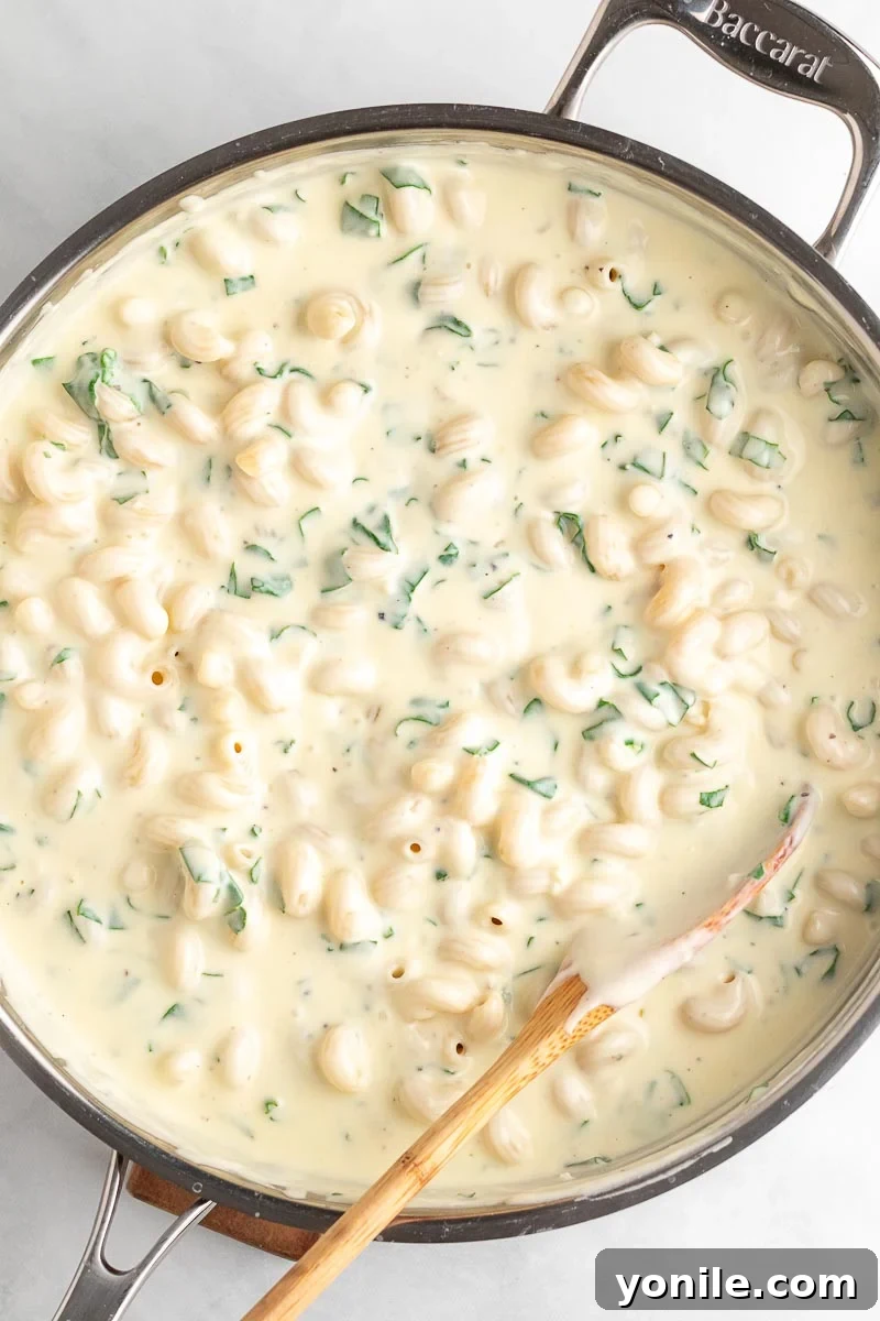 Velvety Spinach Macaroni and Cheese 7 A close-up shot of creamy spinach mac and cheese in a skillet with a wooden spoon, garnished with fresh herbs, showcasing its rich texture.