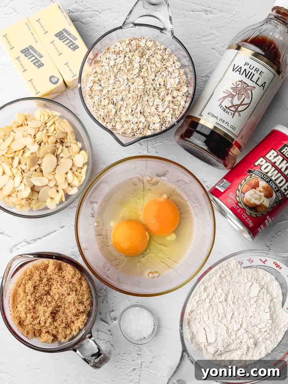 Crispy almond oatmeal cookie ingredients laid out on a wooden surface, including butter, sugar, eggs, vanilla, flour, salt, baking powder, sliced almonds, and quick rolled oats.