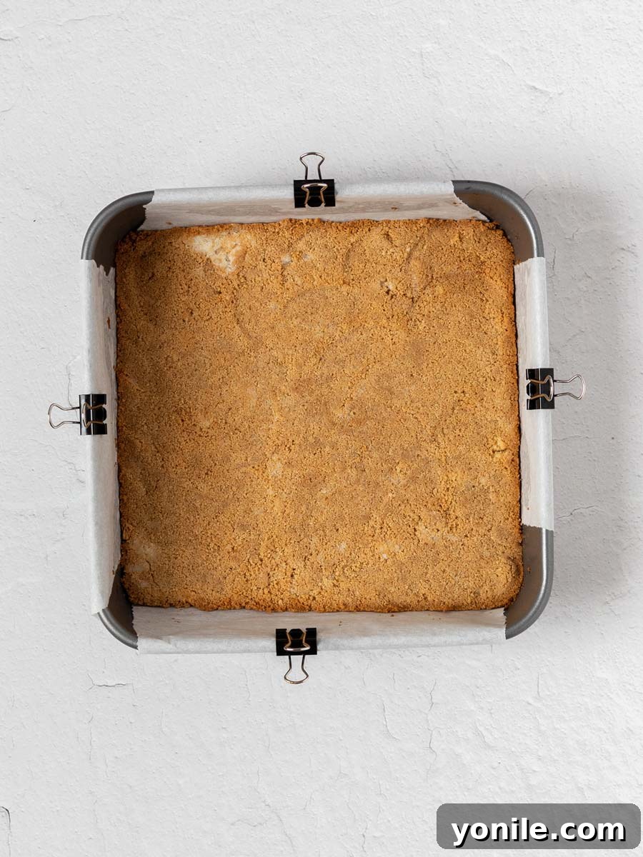 A baked graham cracker crust, golden brown and perfectly pressed into a square baking pan, cooling on a wire rack.