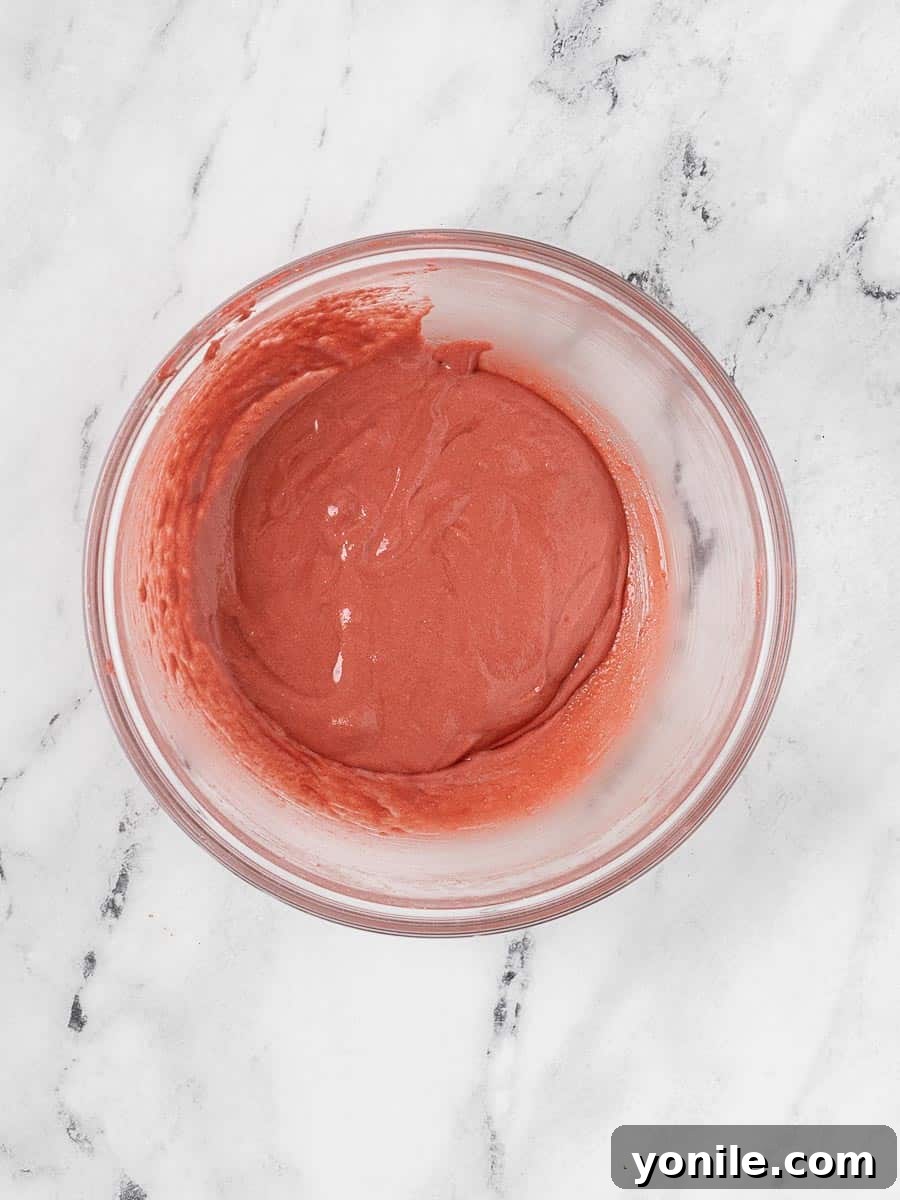 The perfectly mixed macaron batter, a smooth, glossy, and vibrant red, gracefully flowing from a silicone spatula into a bowl, demonstrating ideal macaronage consistency.
