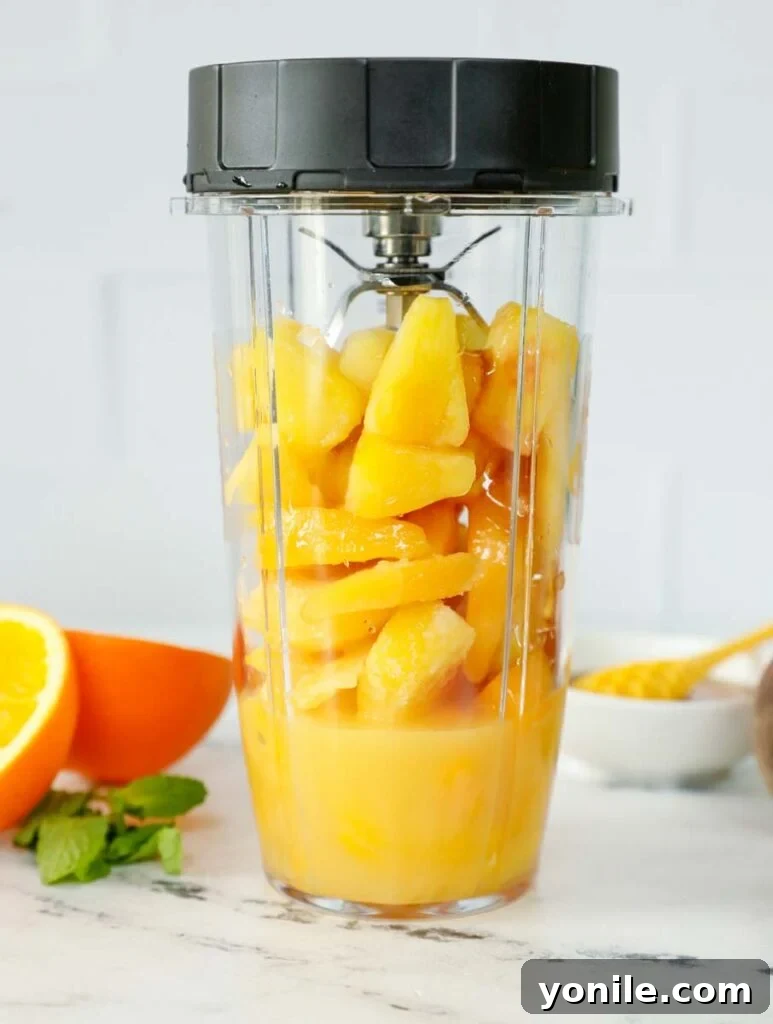 Frozen peach slices and pineapple chunks, along with orange juice, inside a blender, ready to create a delicious Peach N' Orange slushie.