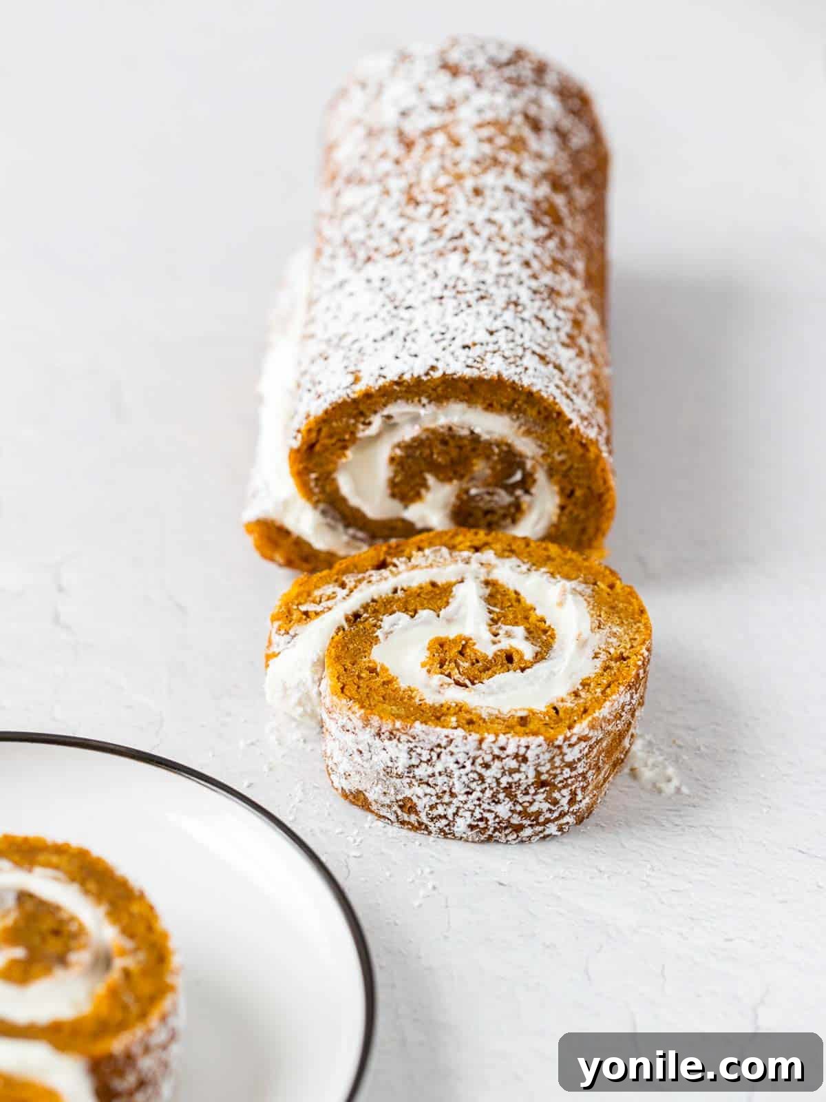 A beautifully sliced pumpkin cake roll revealing its creamy filling, presented alongside the full roll, dusted with powdered sugar, creating an inviting display.