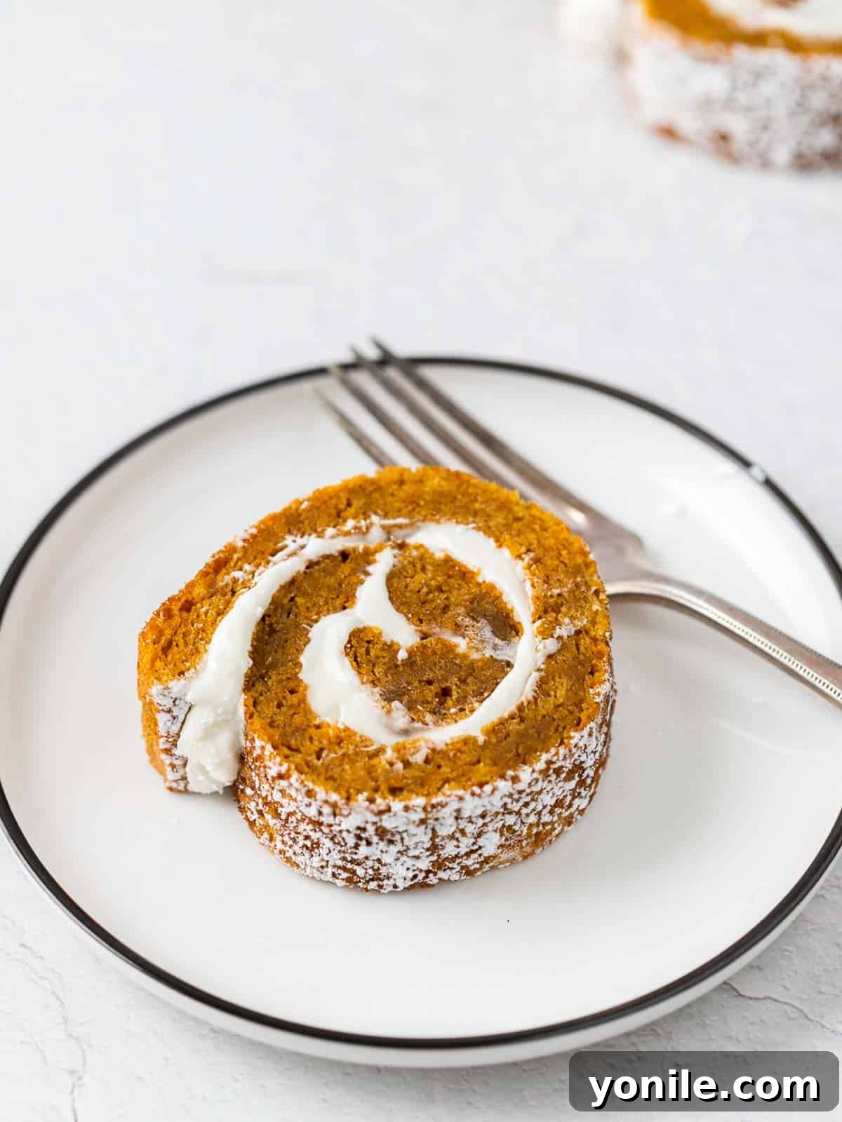 A delectable slice of pumpkin cake roll, showcasing its perfect spiral of cake and cream cheese frosting, elegantly placed on a white plate with a fork.