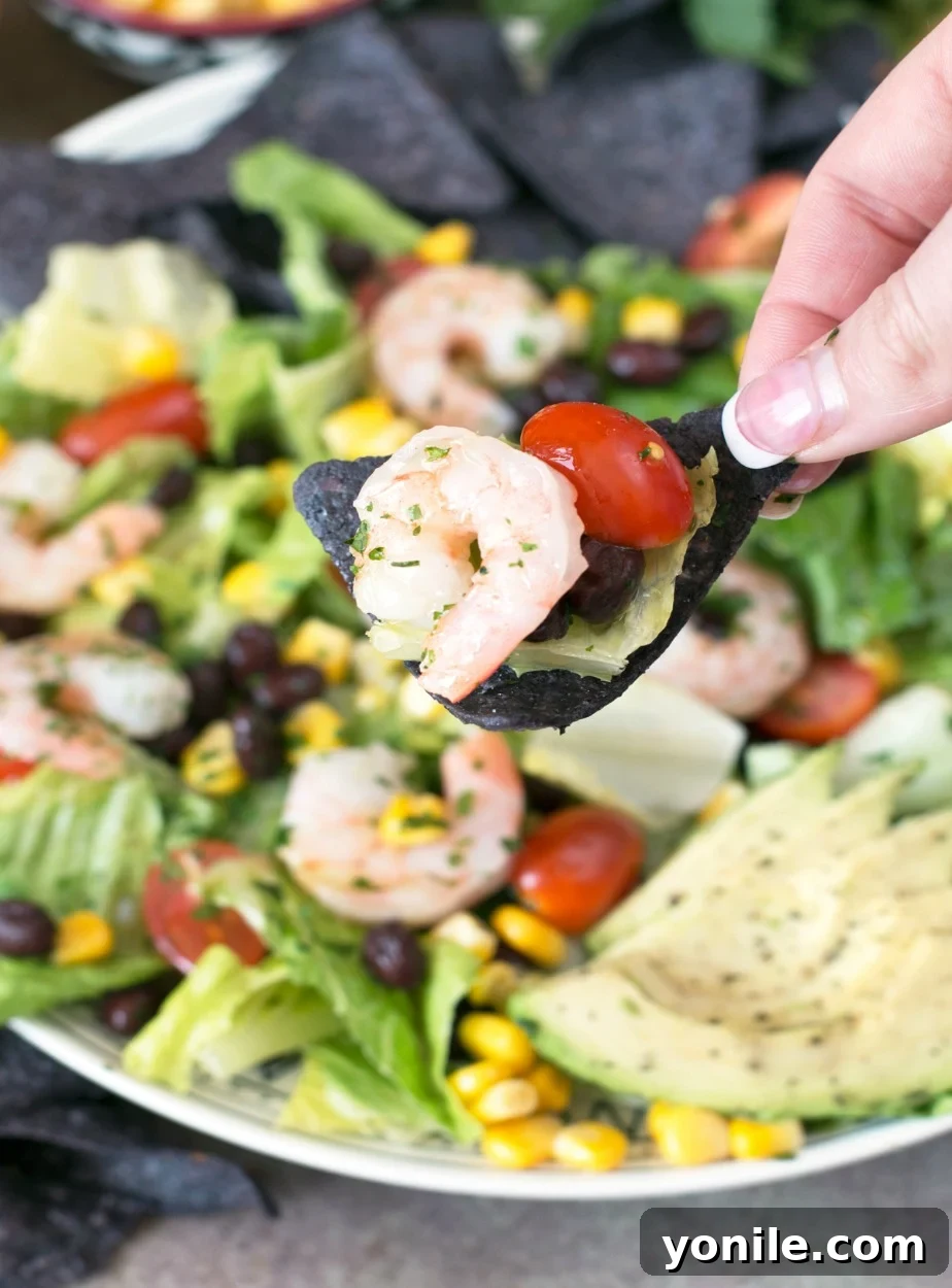A close-up shot of a single bite of Mexican Shrimp Taco Salad, perfectly balanced on a blue corn tortilla chip, held by a woman's hand, ready to be enjoyed.