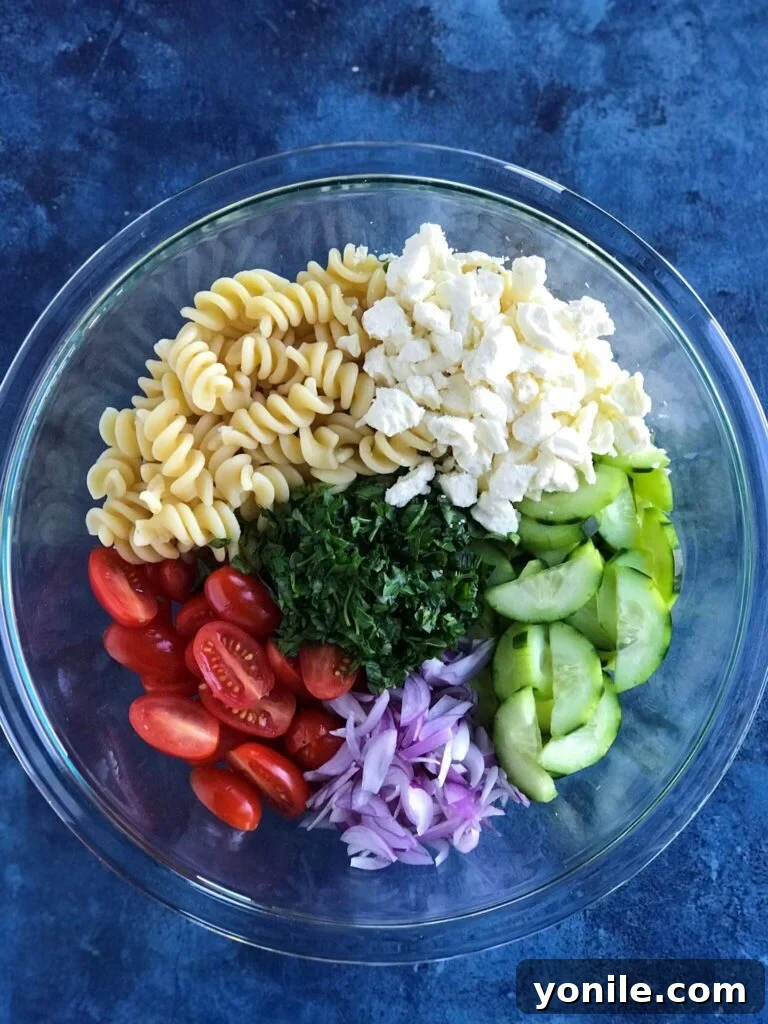 Close-up of Greek Pasta Salad ingredients: feta cheese, rotini pasta, fresh cucumbers, grape tomatoes, red onion, and vibrant fresh parsley, before dressing is added