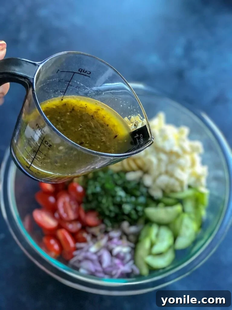 Chef pouring homemade lemon vinaigrette over a bowl of fresh Greek Pasta Salad ingredients, ensuring every piece is coated