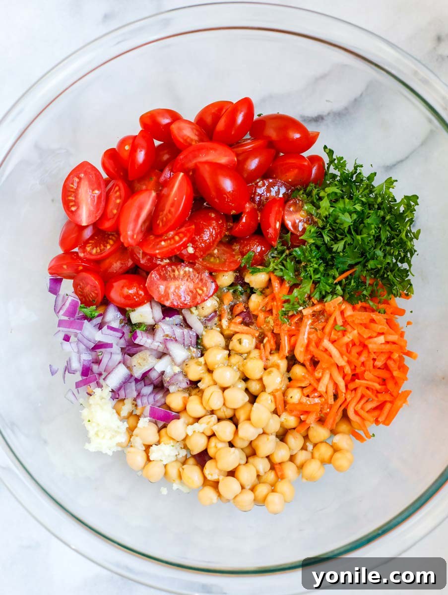 A large bowl filled with various chopped salad ingredients, including tomatoes, herbs, carrots, chickpeas, onions, and garlic, ready for dressing.