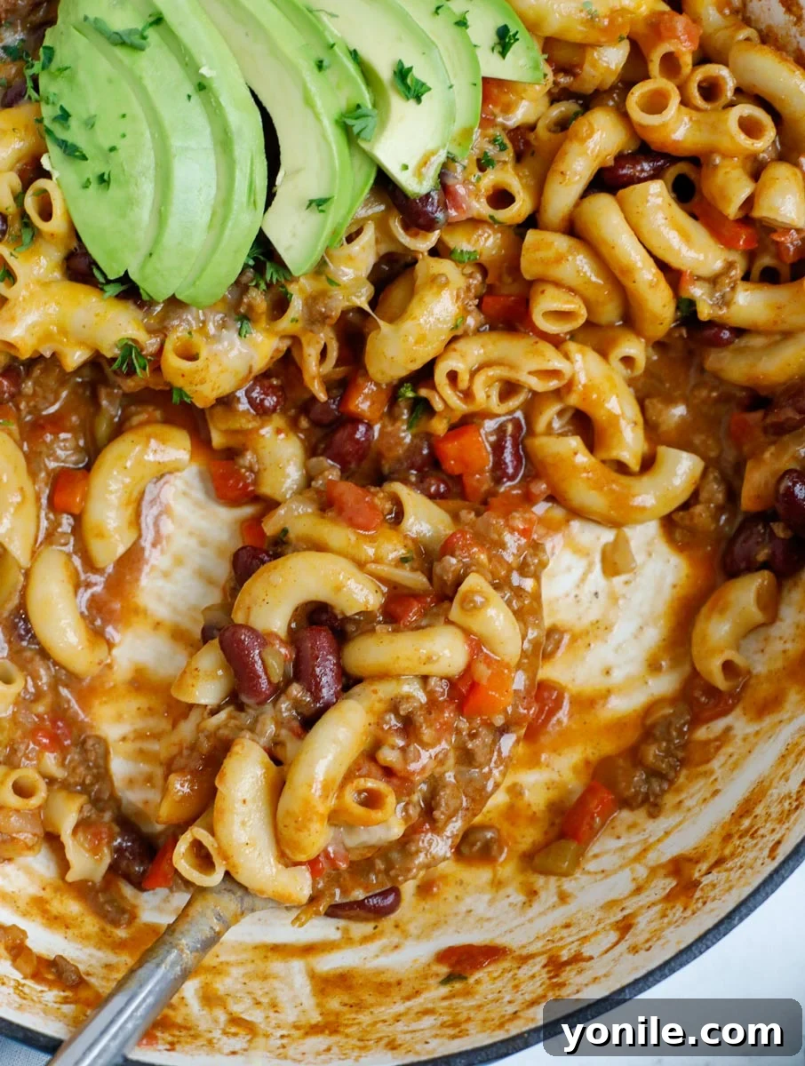 Close up view of a spoon scooping a generous portion of cheesy chili mac from a large pot.
