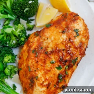 Close up of an air fryer rosemary chicken breast beside some broccoli and lemon wedges.