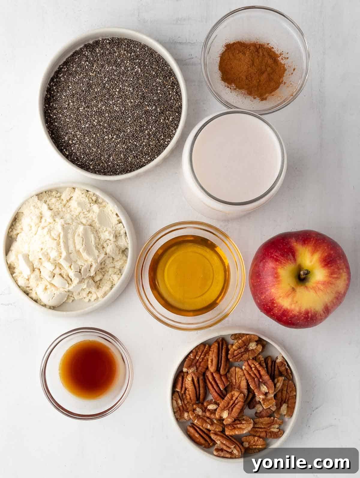 All the fresh ingredients for cinnamon apple overnight chia pudding laid out on a kitchen counter, including chia seeds, protein powder, maple syrup, vanilla extract, almond milk, apples, and cinnamon.