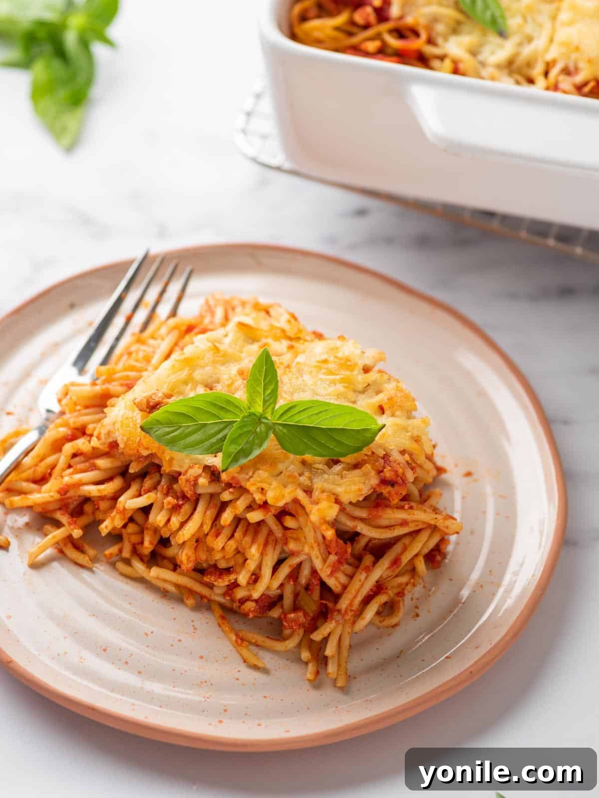Simple Baked Spaghetti Casserole 9 A perfectly portioned slice of baked spaghetti pasta served on a white plate, garnished with fresh basil leaves.