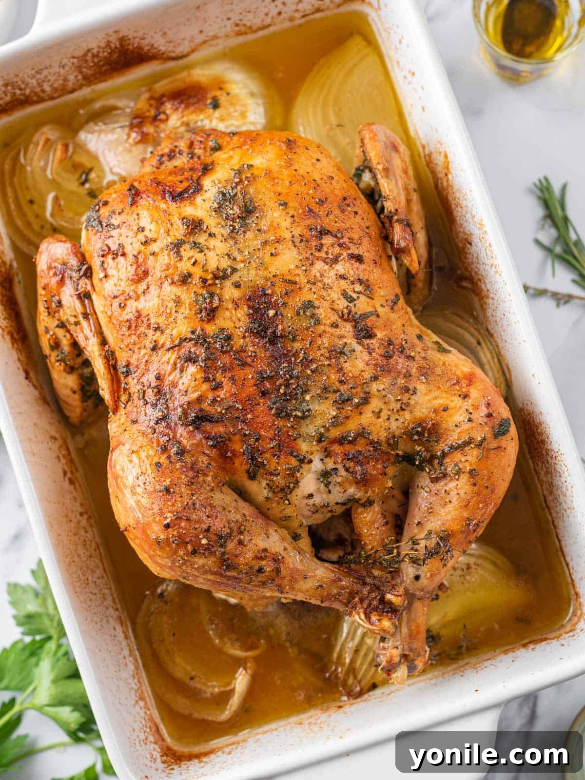 A whole roasted baked chicken in a baking dish, perfectly golden with a rich pan sauce, ready to be removed from the oven.