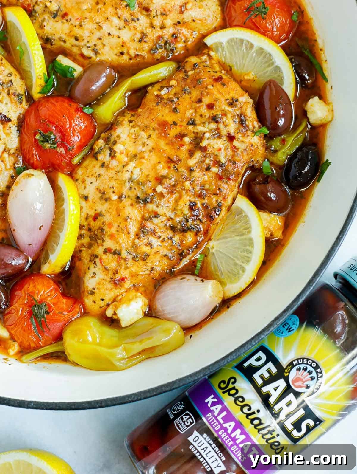 Close-up of a pan of baked Greek chicken with a jar of olives in the background.